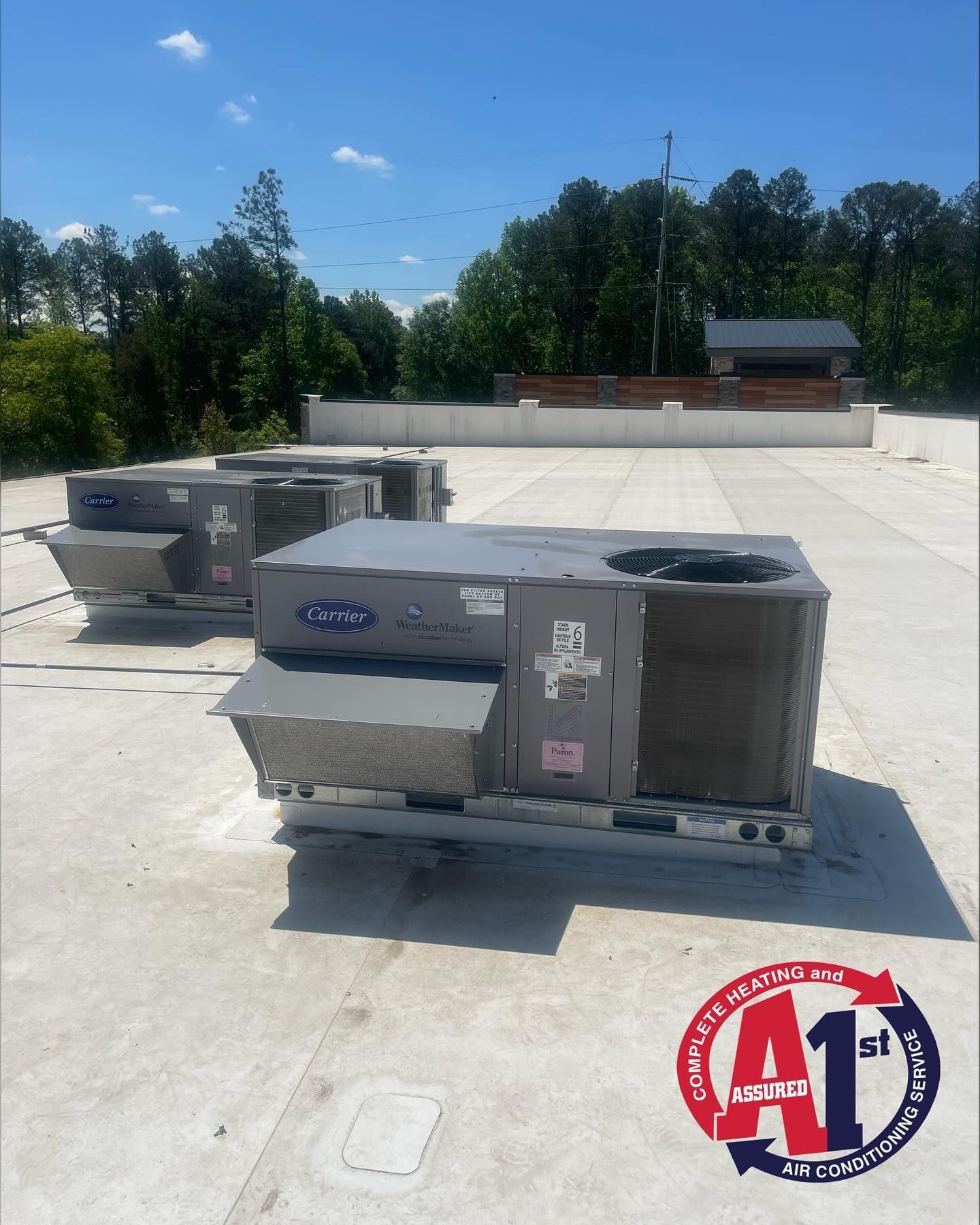 Three rooftop air conditioning units on a flat, white roof under a blue sky, trees in the background.