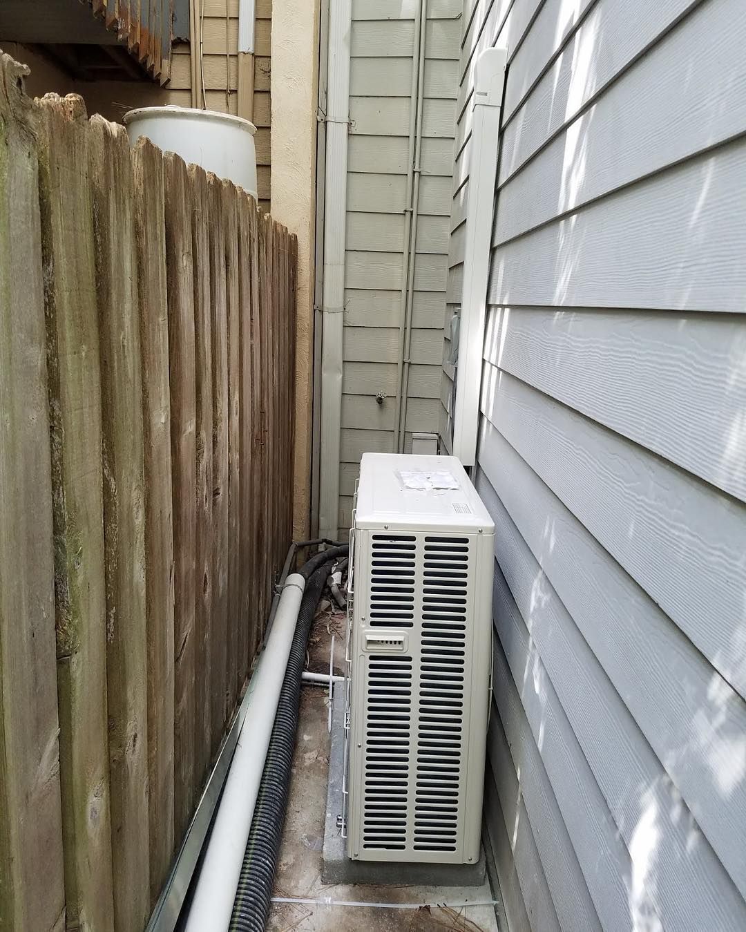 Air conditioning unit between a wooden fence and a house with light grey siding.