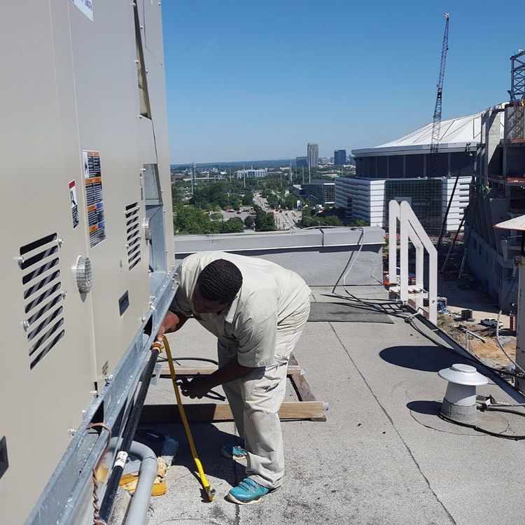 Person in tan work clothes measures on a rooftop next to HVAC equipment with a cityscape in the background.