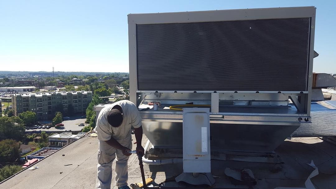 A person works on a rooftop cooling system with city buildings in the background. Sunny day.