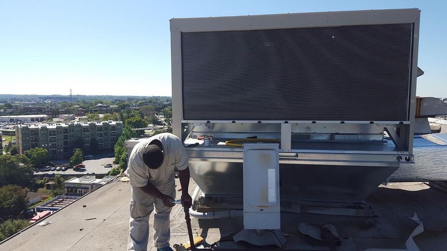 A person works on a rooftop cooling system with city buildings in the background. Sunny day.