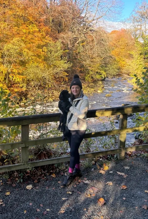 Julie holding a small black dog stands on a path by a river with autumn foliage.