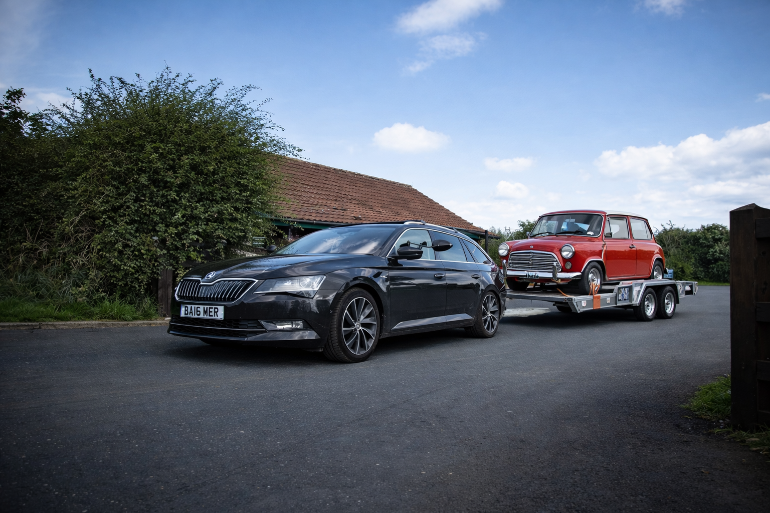 A dark gray car towing a trailer with a classic red car, set outdoors with a house in the background.
