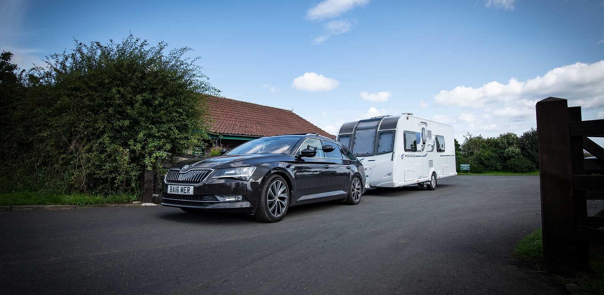A black car towing a white caravan on a paved road. Blue sky, a building, and trees in the background.