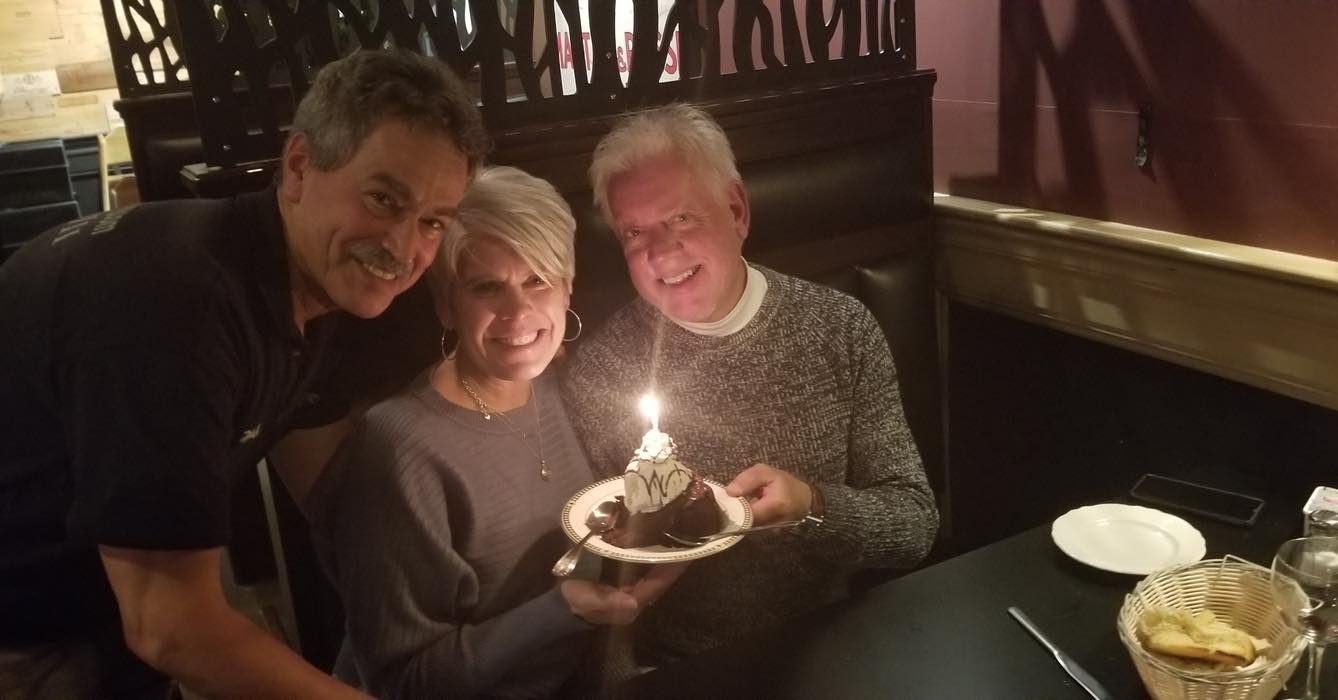 Two men and a woman are sitting at a table holding a cake with a candle on it