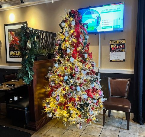 A decorated Christmas tree in a restaurant with a television, a framed poster, and a chair.