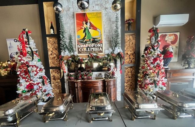 Buffet setup with holiday decor; silver serving dishes on tables. Christmas trees, poster, fireplace backdrop.