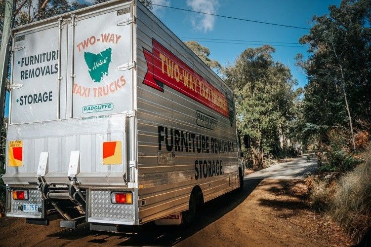 A moving truck travels down a dusty dirt road surrounded by trees and open fields.