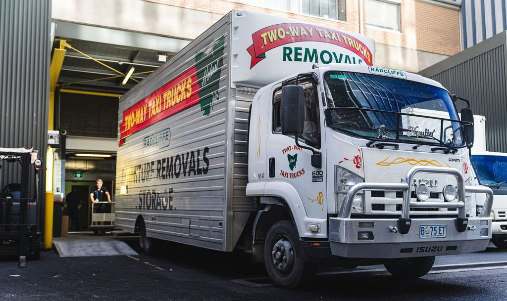 Two movers in red uniforms carry a white bed frame against a plain white background