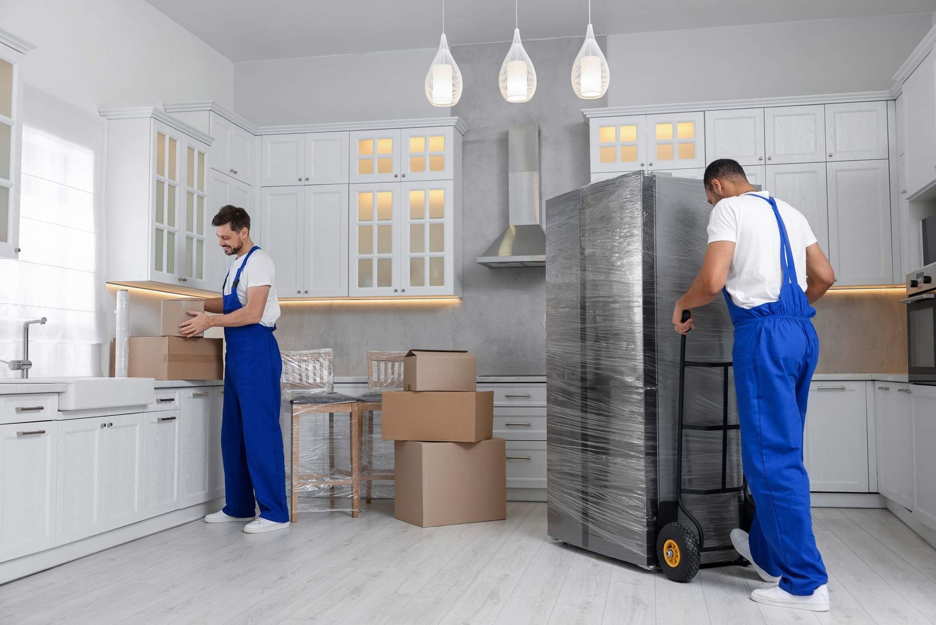 Two workers in blue overalls moving boxes and a wrapped fridge in kitchen. Two workers in blue overalls moving boxes and a wrapped fridge in kitchen.