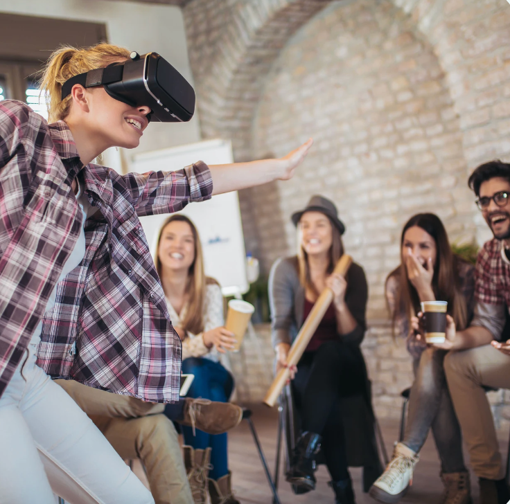 Woman in VR headset gestures wildly as friends watch, laughing. They sit in a circle in a brightly lit room with arches.
