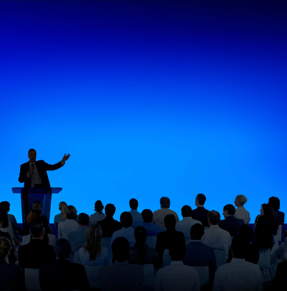 A man giving a presentation at a podium to an audience in front of a blue screen.