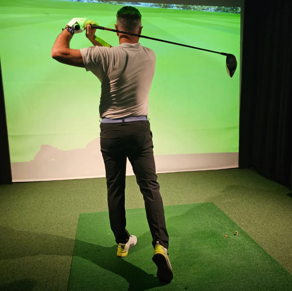 Man in a golf simulator, mid-swing with a club. He's wearing grey shirt and black pants, striking the ball on a green mat.