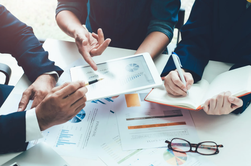 People in business attire collaborating around a table, examining financial reports and a tablet, with pens and glasses.
