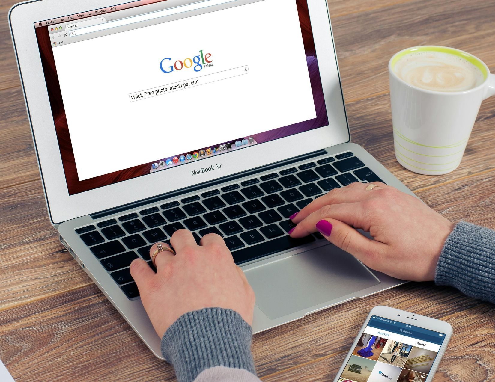 Person typing on a laptop displaying the Google homepage, with a smartphone and mug of coffee on a wooden table.
