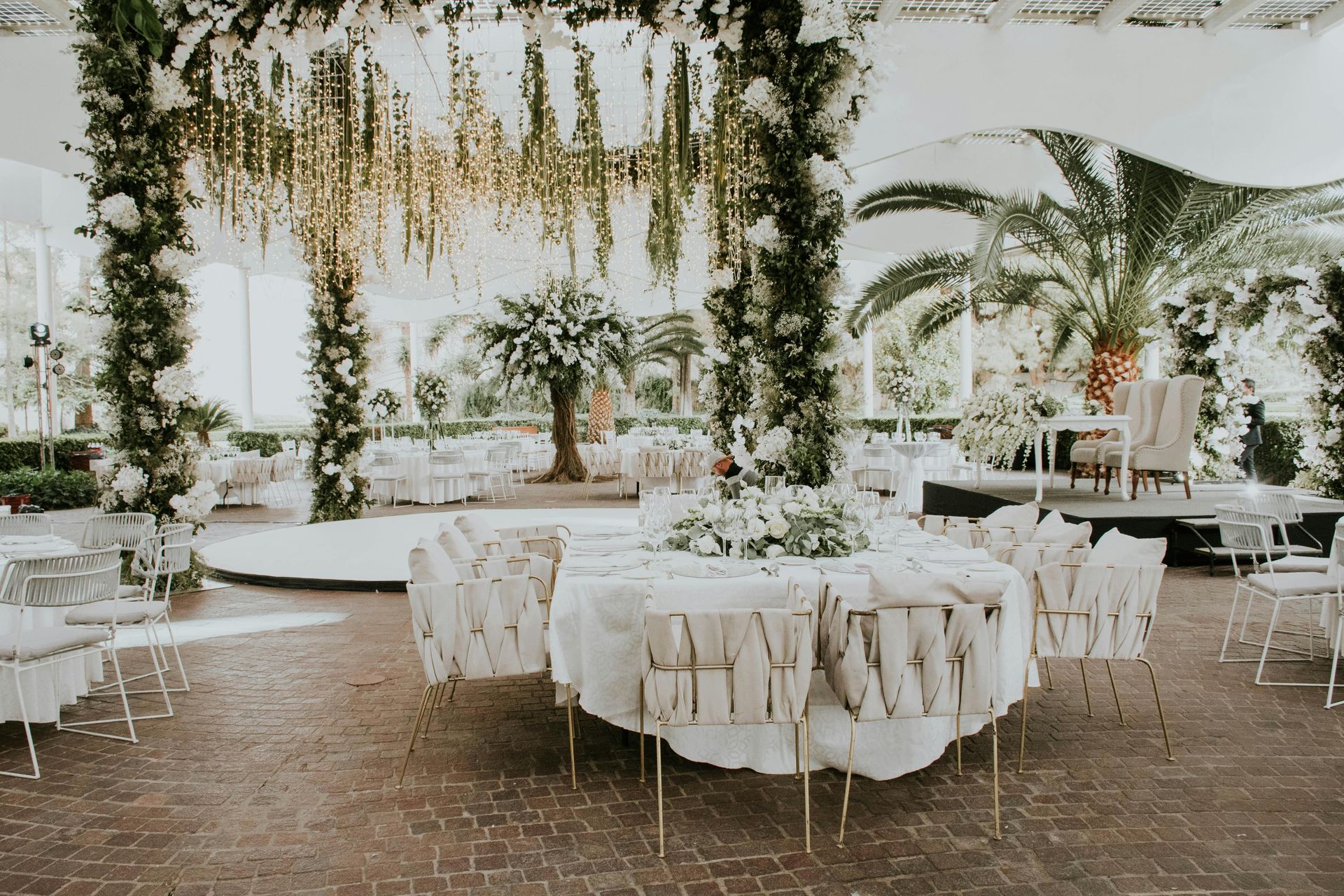 Wedding reception setup with white floral arrangements, a round table, and elegant white chairs, outdoors.