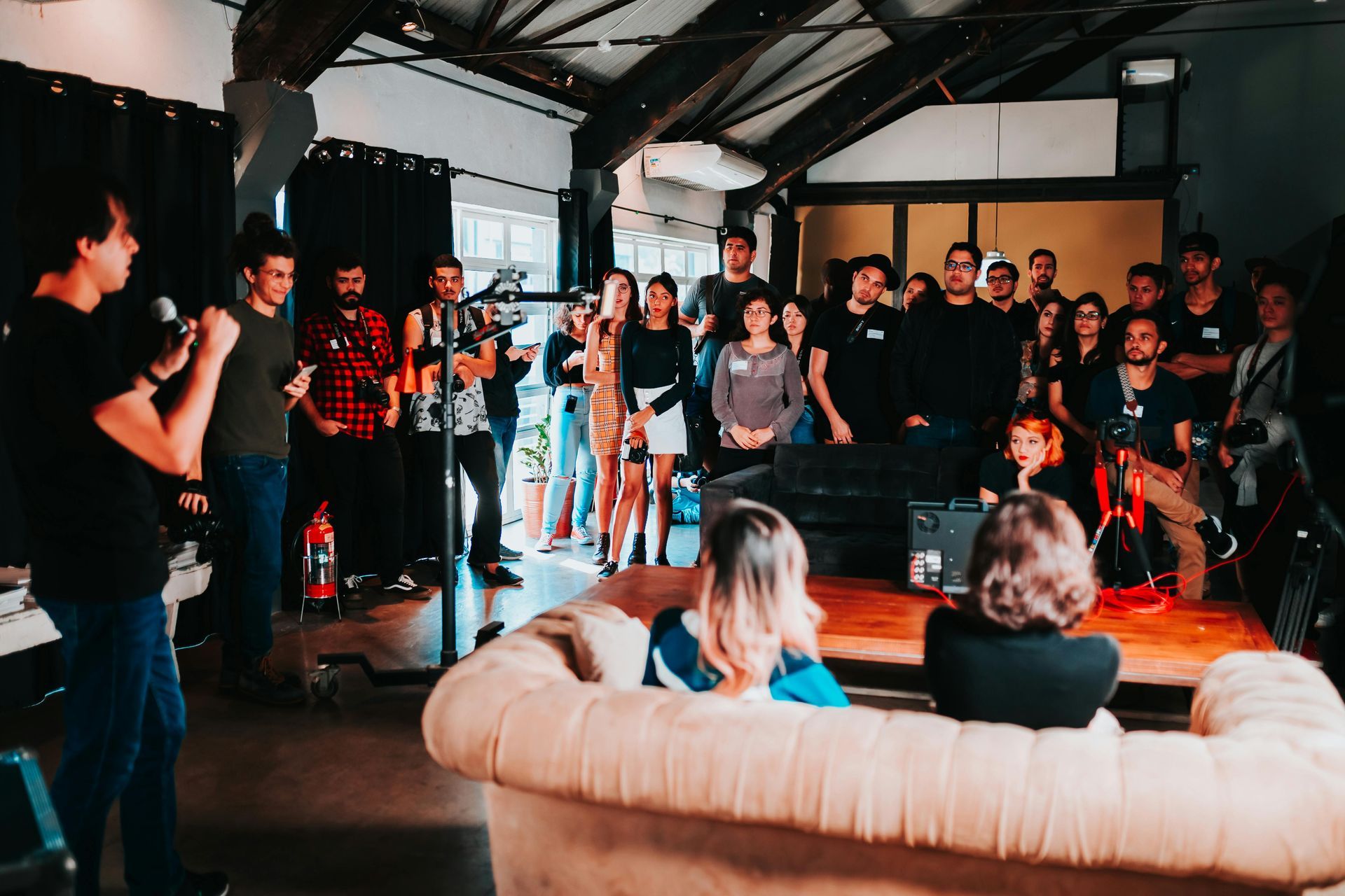 A presenter speaks at a gathering in a loft space. A crowd of people stands, some listening, others looking at a screen.