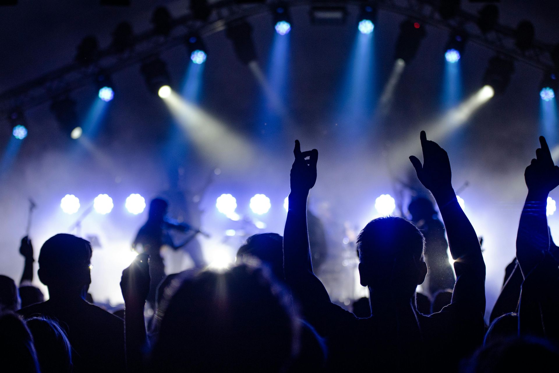 Concert crowd with raised arms silhouetted against a brightly lit stage with musicians. Blue stage lighting.