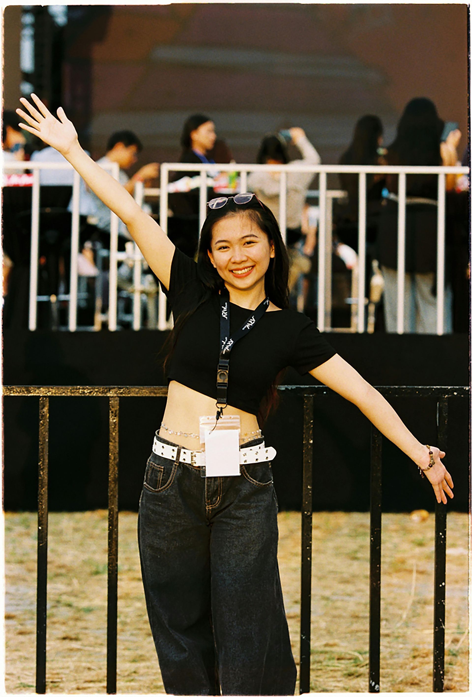 Young woman with arms raised, smiling, wearing black crop top, jeans, and a white belt outdoors in front of a stage.