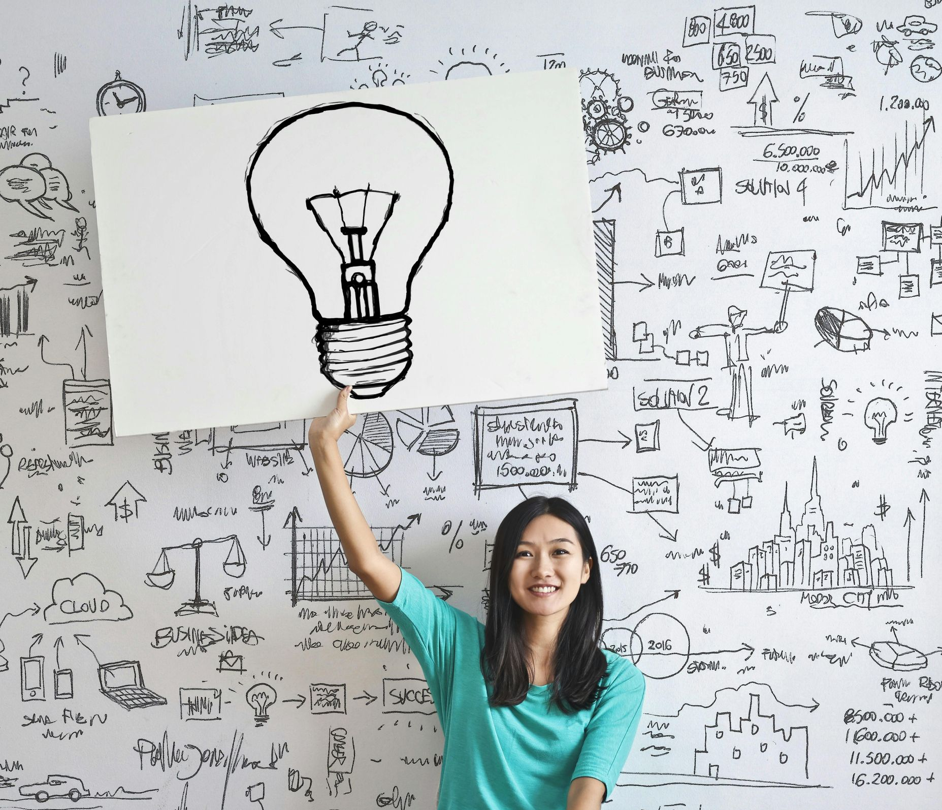 Woman holding a sign with a lightbulb sketch, standing in front of a wall covered in strategy planning doodles. She smiles.