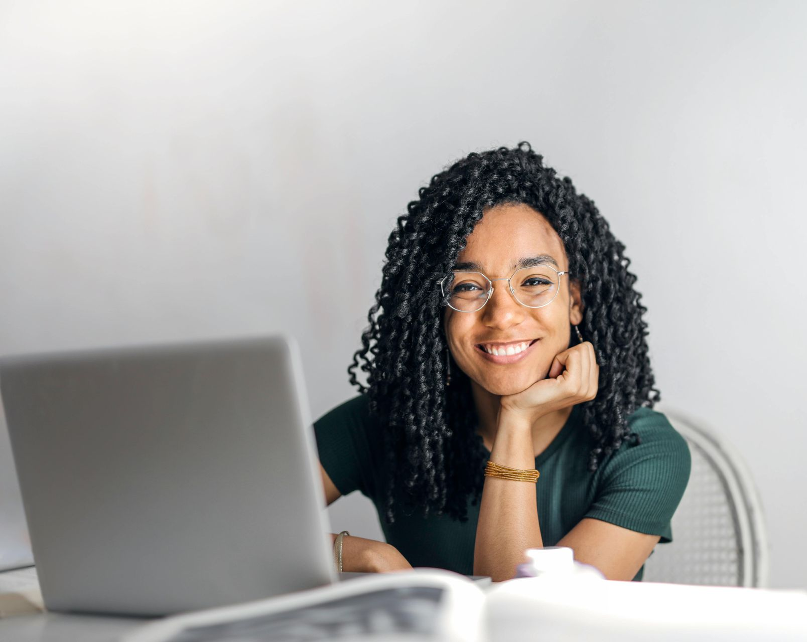 A smiling woman with glasses sits at a desk, using a laptop. She rests her chin on her hand, looking directly at the camera.
