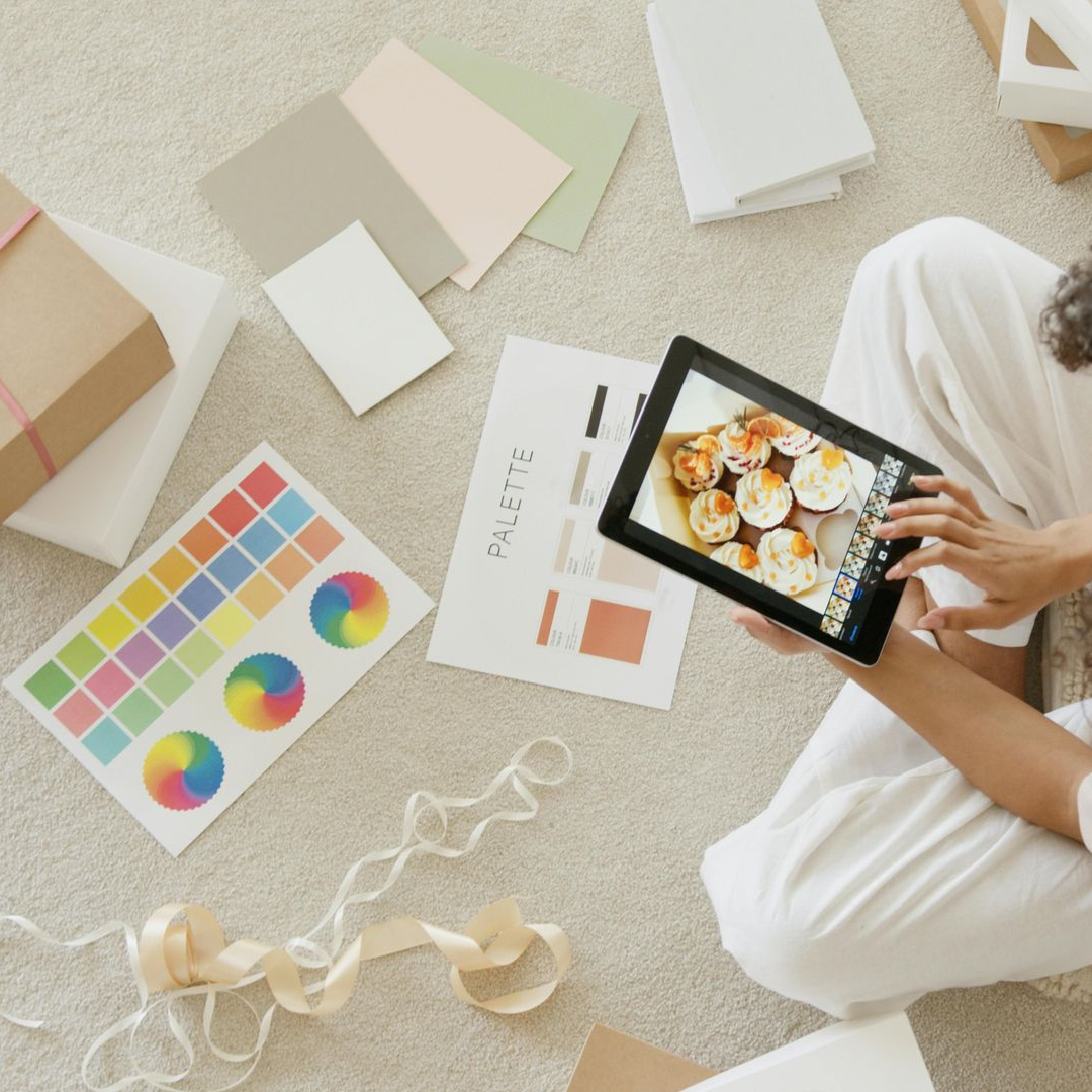 Person seated, holding a tablet. Surrounded by design materials: color palettes, gifts, and ribbon.