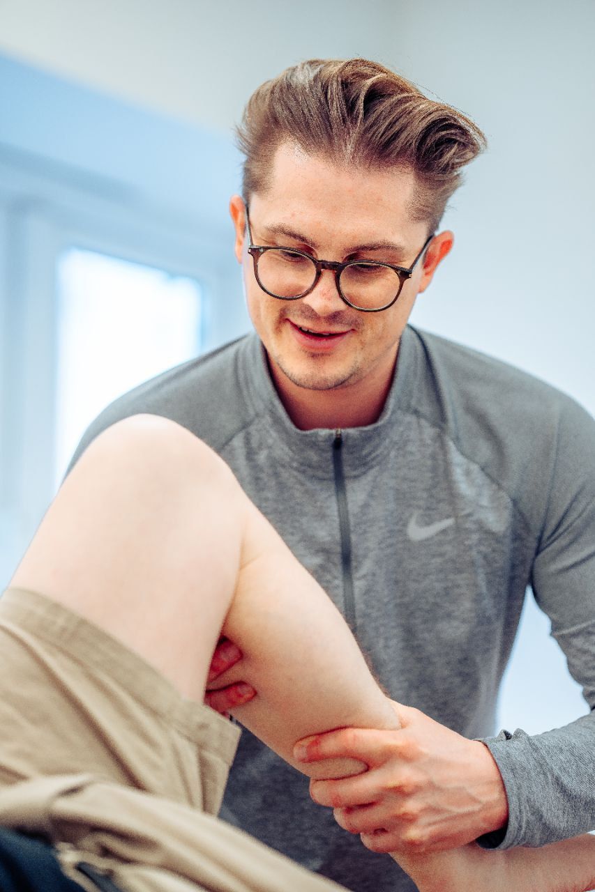 A man is helping a woman stretch her legs on a yoga mat in a gym.