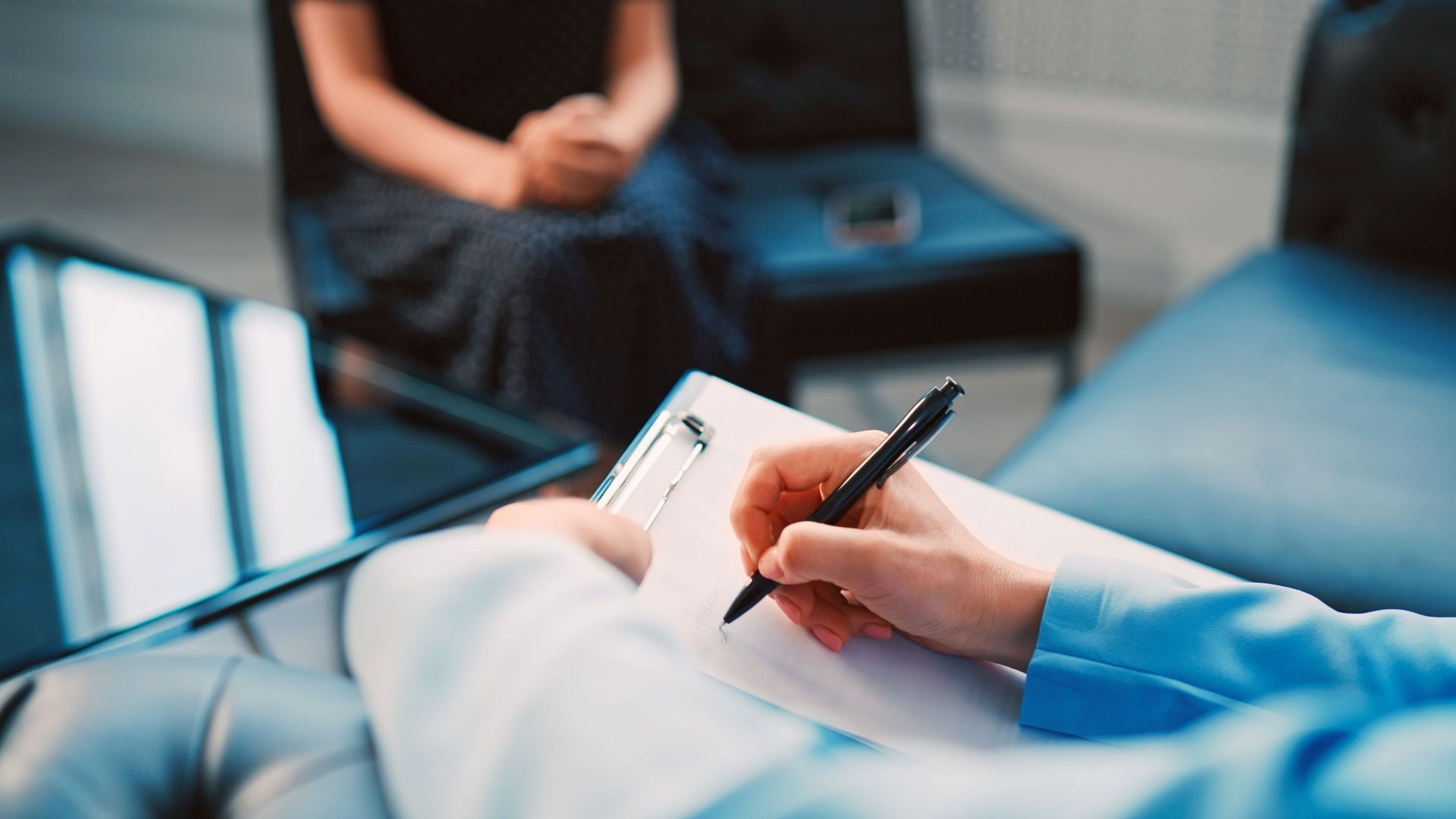 A person is writing on a clipboard while a woman sits in the background.