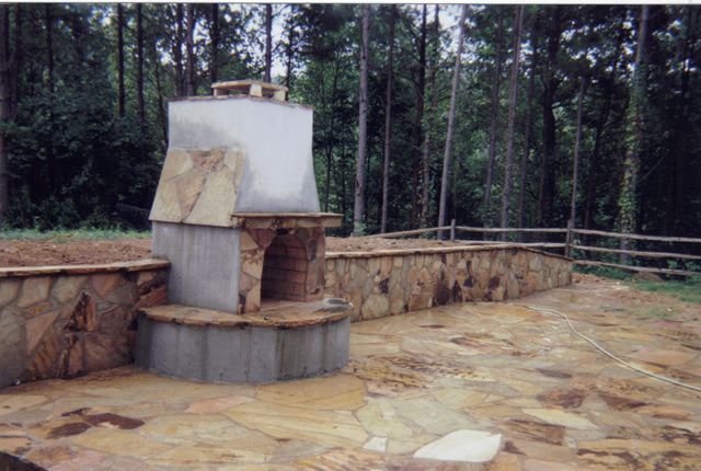 There is a fireplace in the middle of a stone patio surrounded by trees.