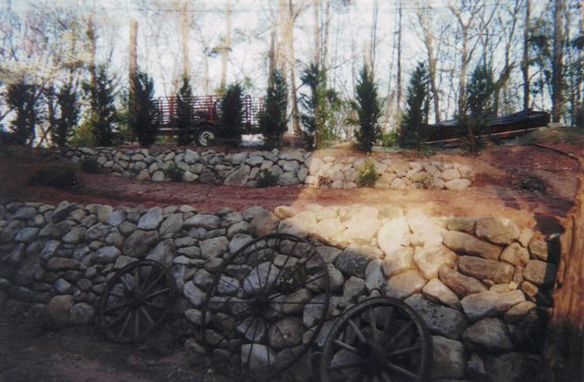 A stone wall with a wagon wheel in the foreground and trees in the background.