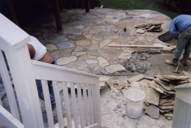 Two men are working on a stone patio with a white railing