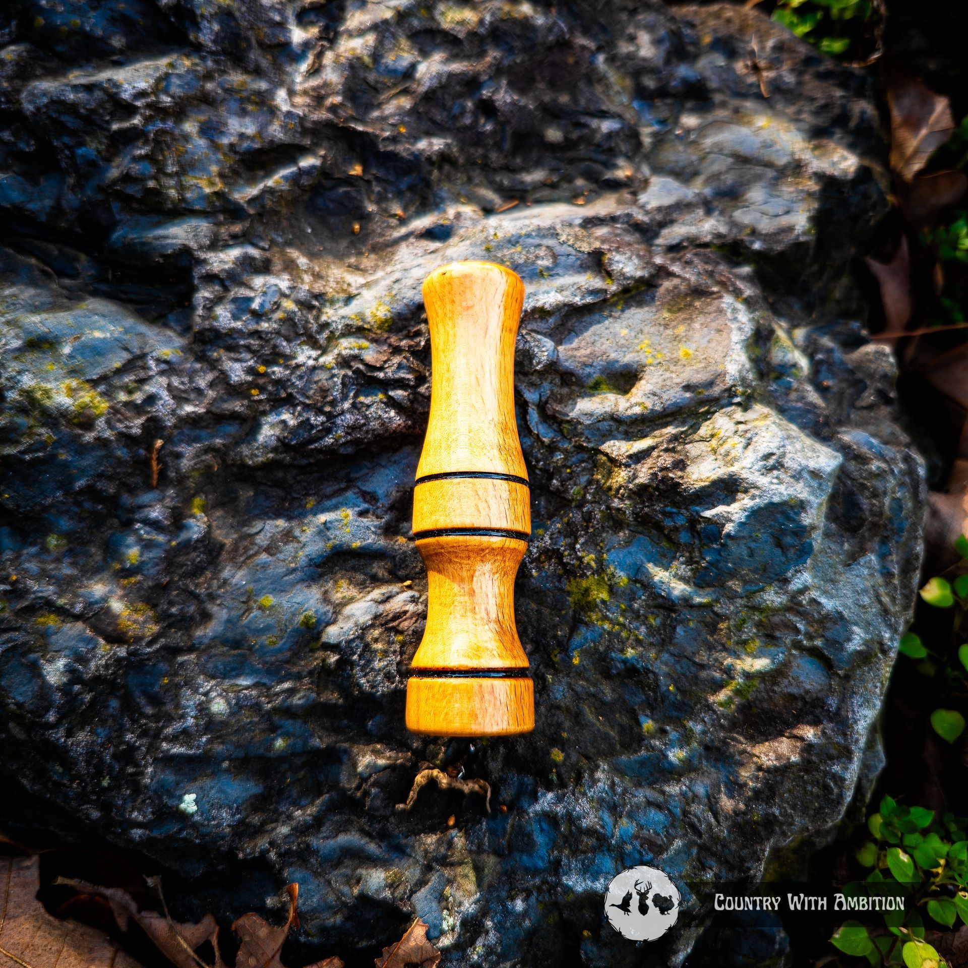 Wooden hand turned predator call on a dark rock, surrounded by leaves. Country with Ambition