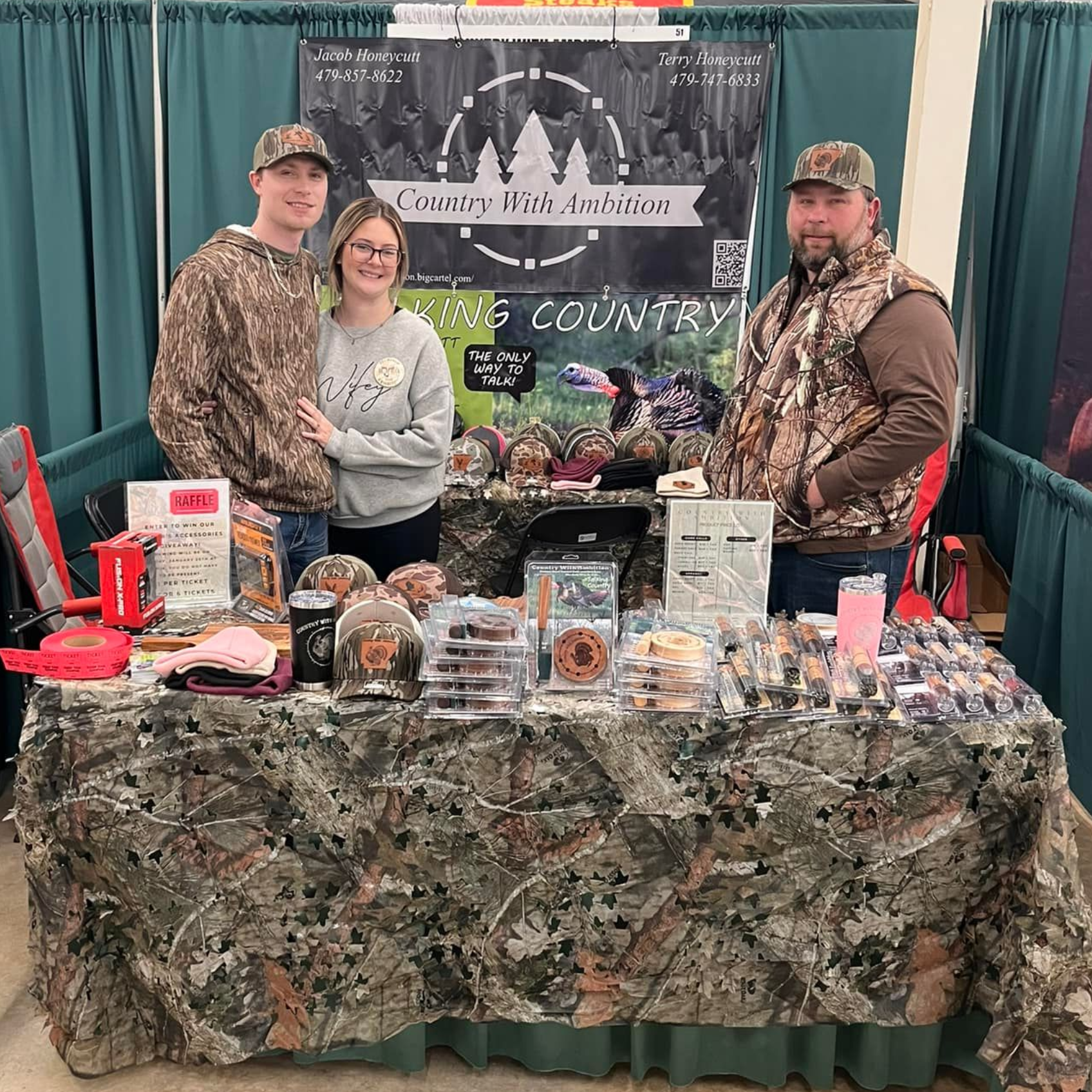 Three people stand behind a booth displaying Country With Ambition merchandise. 
