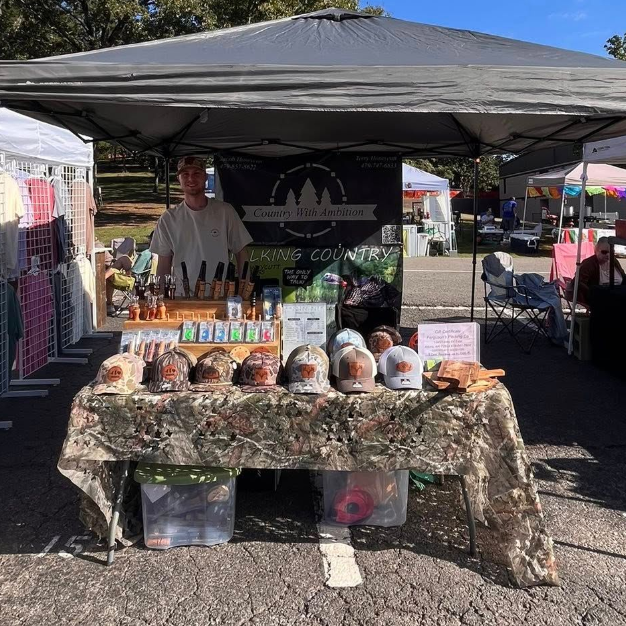 Vendor at an outdoor market under a canopy selling Country With Ambition goods.