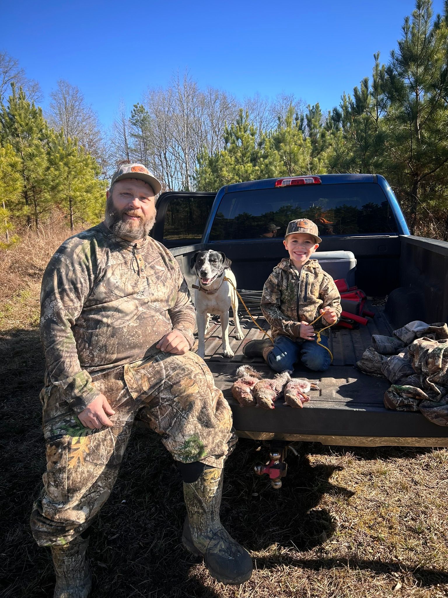 Man and child in camo with dog in truck bed after hunting wearing Country With Ambition Apparel 