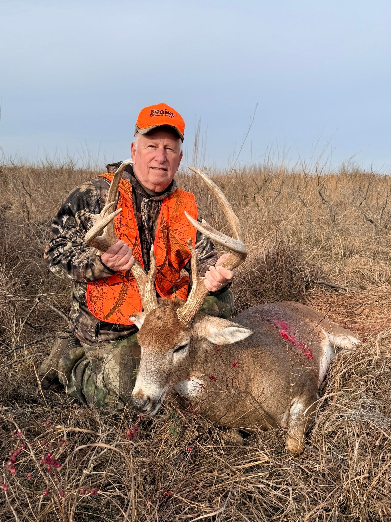 Hunter kneels next to a harvested deer in a field, wearing orange vest and hat. Country With Ambition deer grunt