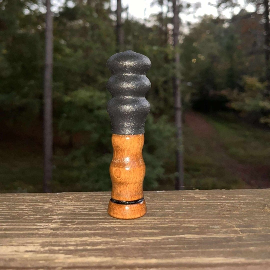 Carved wooden squirrel call on a dark rock, surrounded by leaves and green plants. Country With Ambition