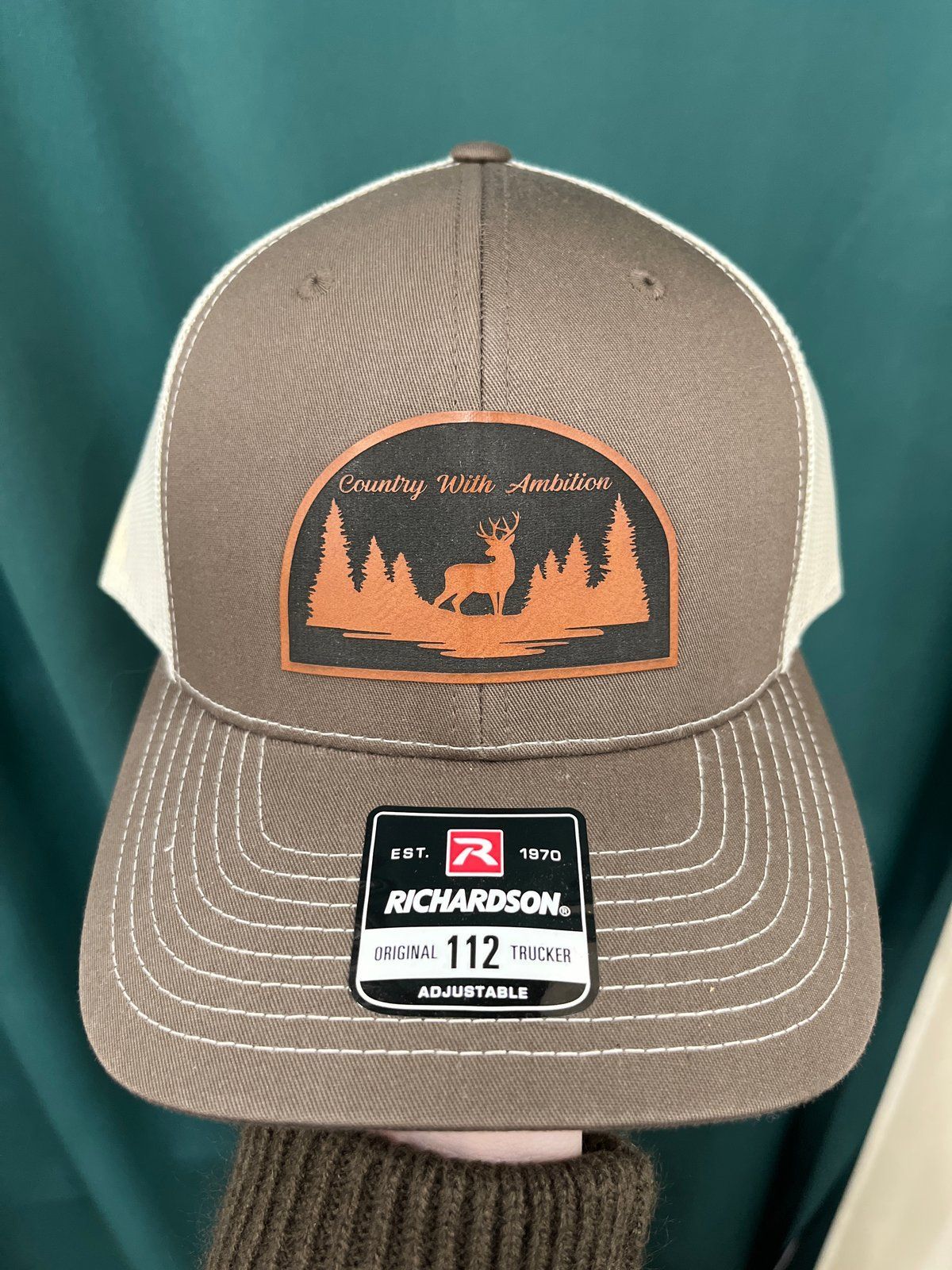 Brown and tan trucker hat with a patch of a deer and trees; Richardson brand. Country With Ambition