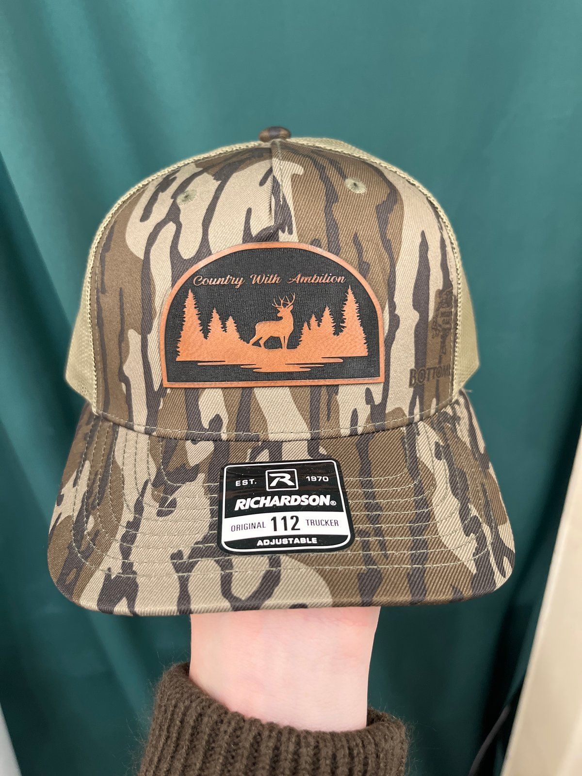 Brown and tan trucker hat with a patch of a deer and trees; Richardson brand. Country With Ambition
