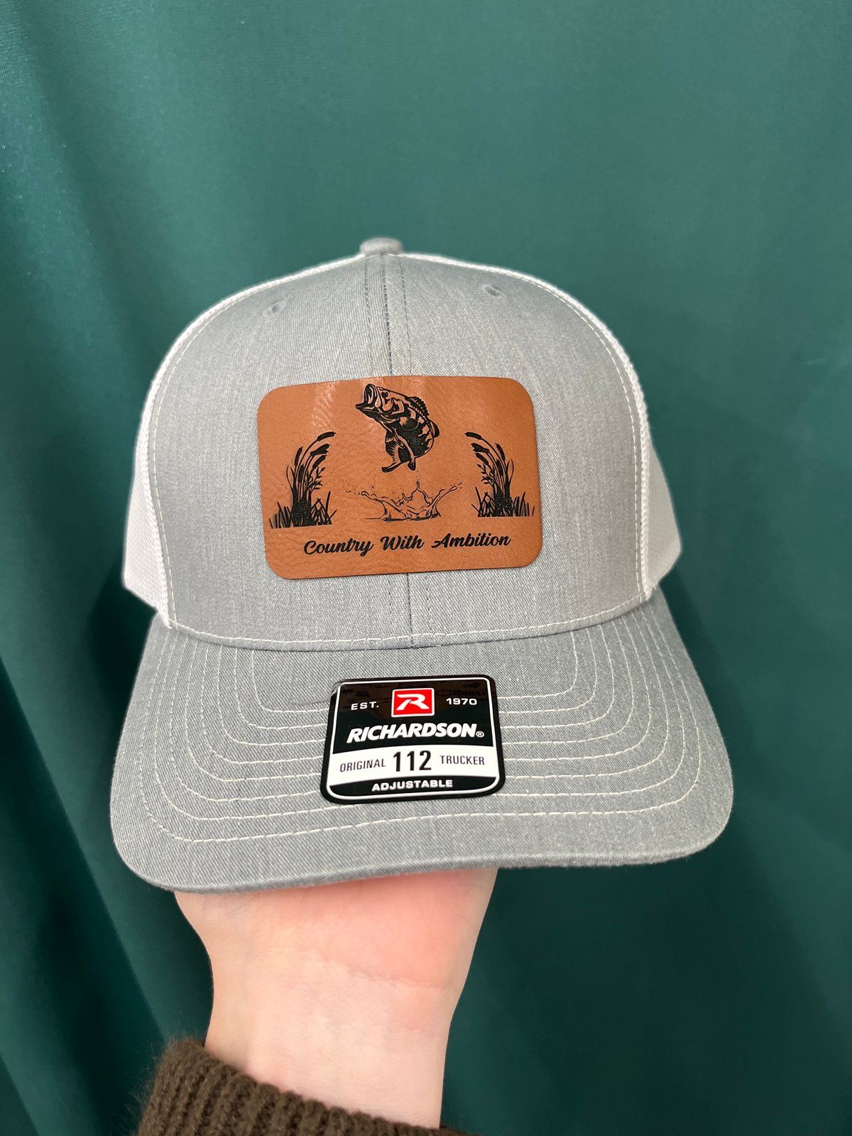 Brown and white trucker hat with a leather patch featuring bass; Richardson Country With Ambition