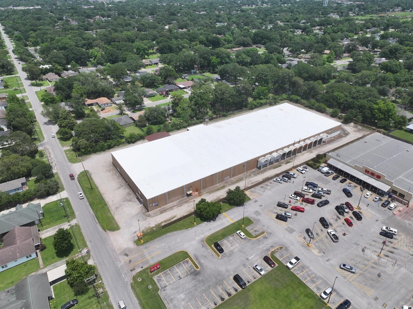 Aerial view of a large warehouse-like building with a white roof and a parking lot full of cars.