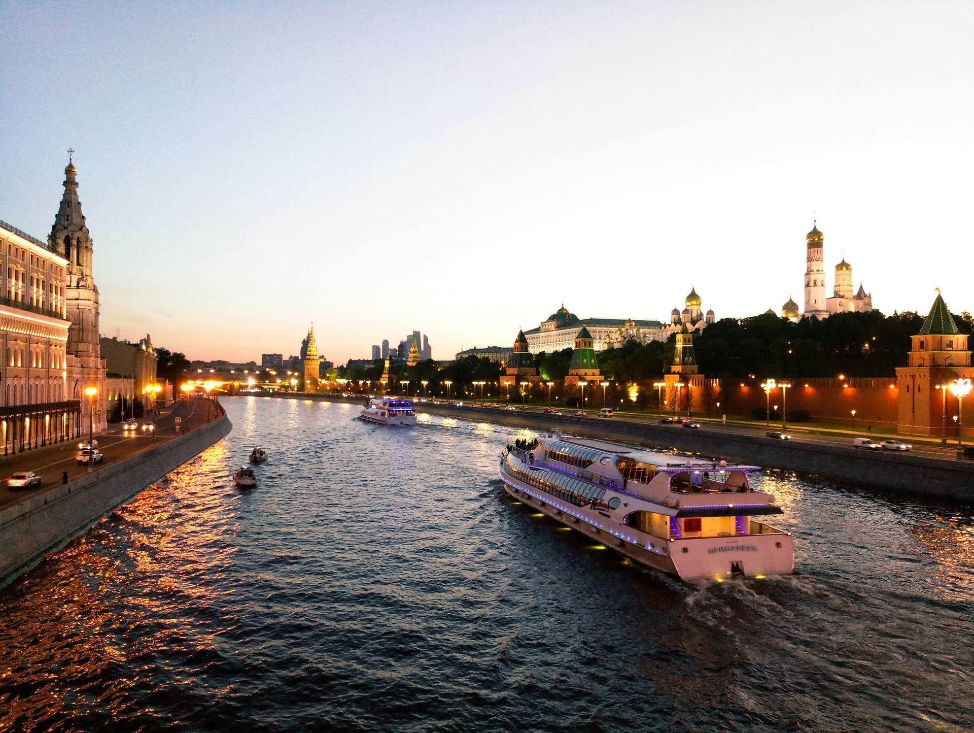 Boats on Moscow River at dusk, with illuminated buildings, including the Kremlin.