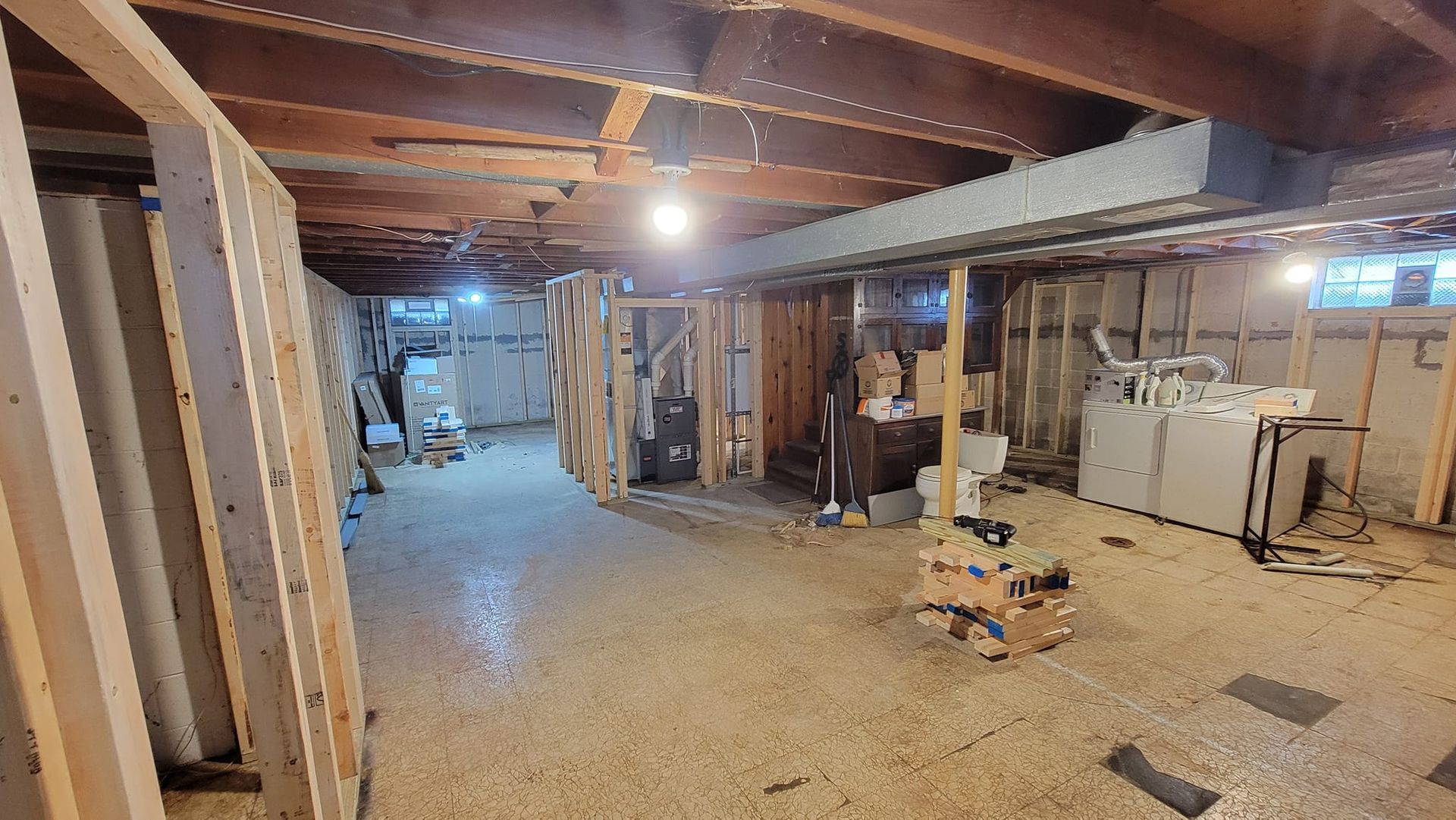 An unfinished basement featuring exposed ceiling joists, wooden framing for walls, a gray duct, and scattered construction.