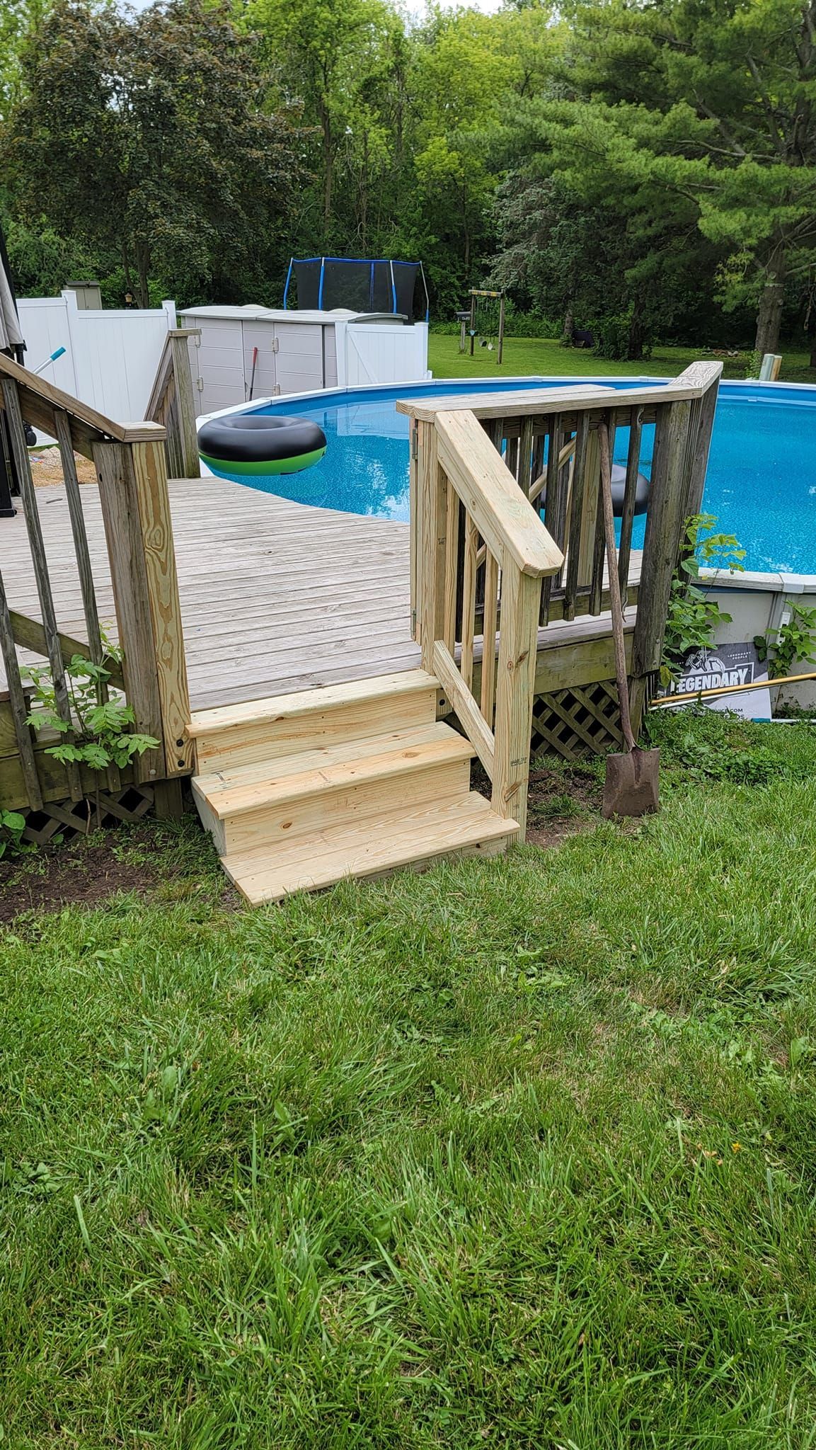 A wooden staircase with railings leads up to a deck beside an above-ground swimming pool in a grassy backyard.