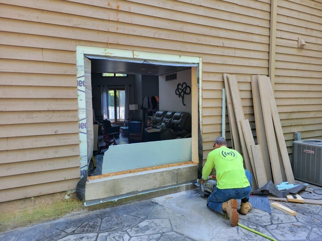 A construction worker in a neon green shirt kneels on a stone patio while cutting into the exterior wall of a house.