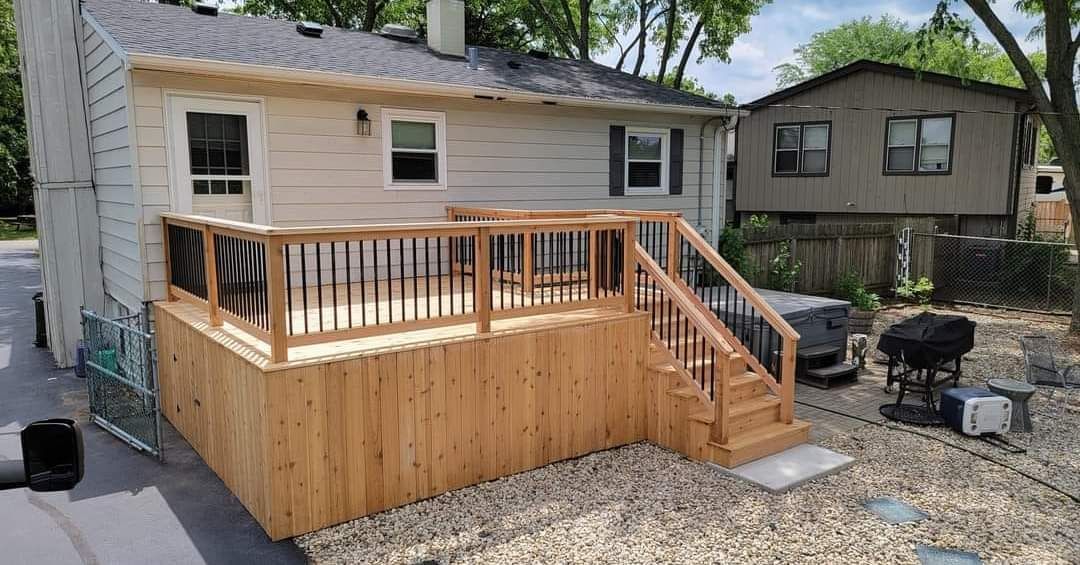 Newly built wooden deck with black railings and stairs, attached to the back of a light-colored house in a backyard.