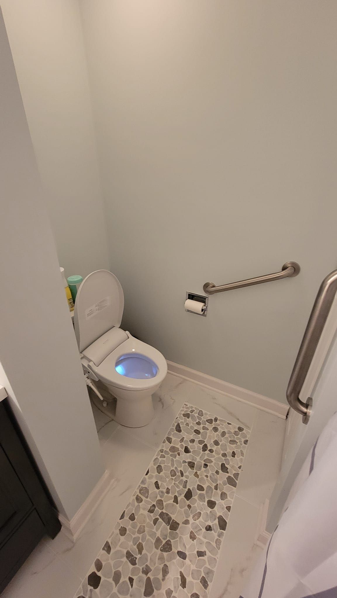 A bathroom stall with a toilet featuring a glowing blue light, a metal grab bar, and a stone-patterned floor runner.