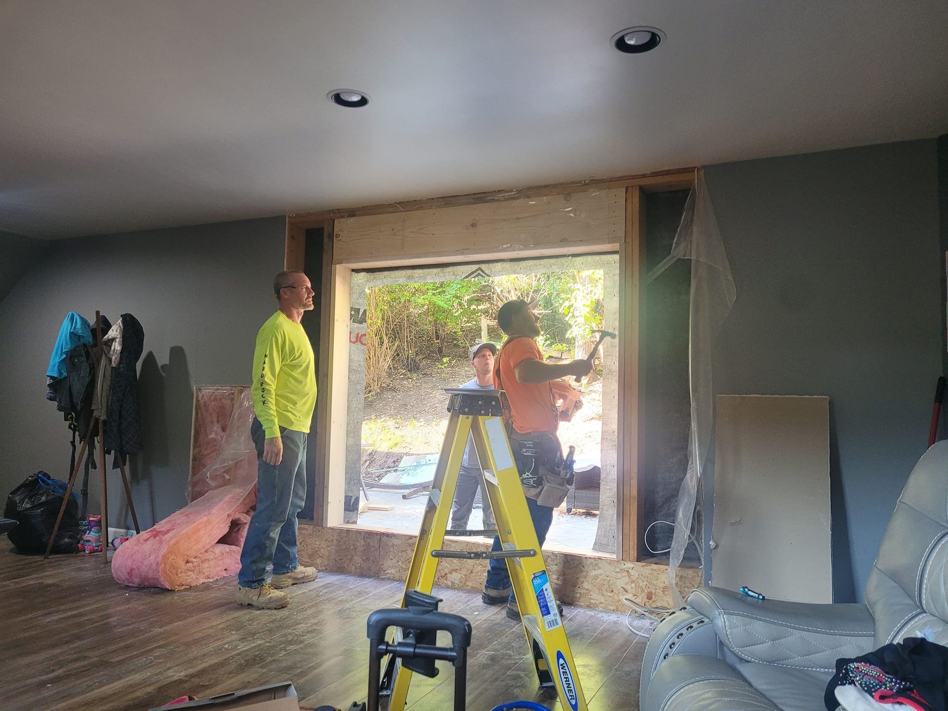 Two construction workers in high-visibility gear work on a large window installation in an indoor room.