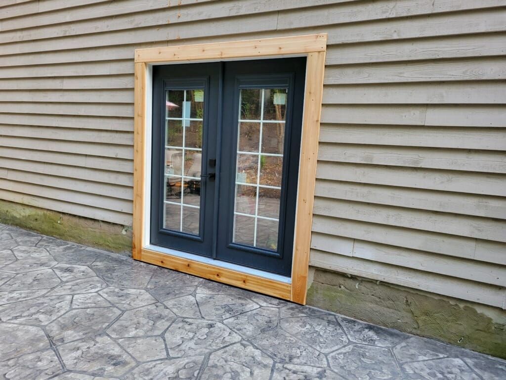 Double French doors with a dark frame and glass panes, installed in a light-sided home with raw wood trim, on a patio.