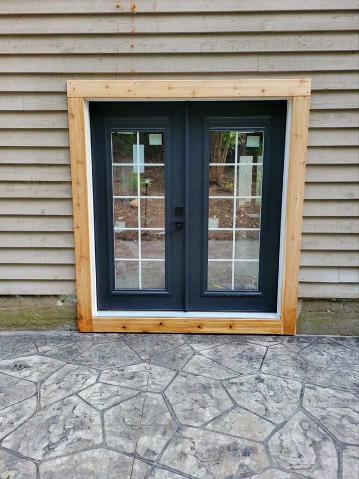 A set of dark-framed double glass doors with white grilles, framed in natural wood against light brown horizontal siding.